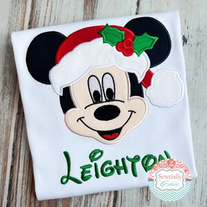 May include: White t-shirt featuring a festive Mickey Mouse design. Mickey is wearing a Santa hat with holly accents. The name "Leighton" is embroidered in green script. A decorative logo is in the bottom right corner.