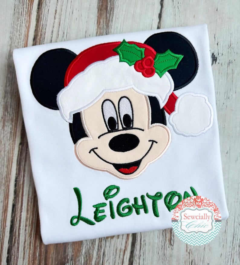 May include: White t-shirt featuring a festive Mickey Mouse design. Mickey is wearing a Santa hat with holly accents. The name "Leighton" is embroidered in green script. A decorative logo is in the bottom right corner.