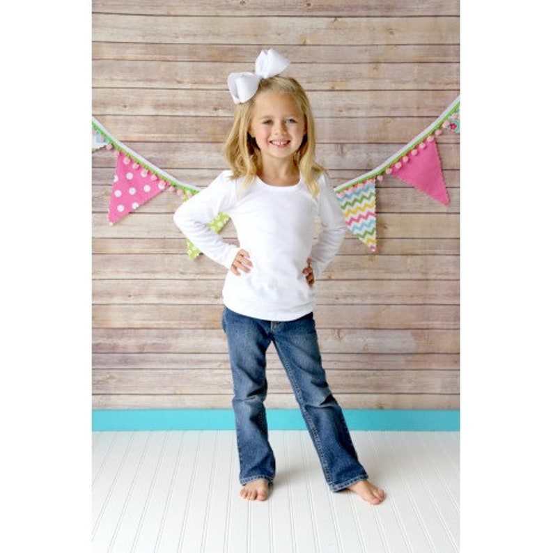 May include: A young girl wearing a white long-sleeved shirt and blue jeans. She is standing in front of a wooden background with a colorful banner hanging above her.