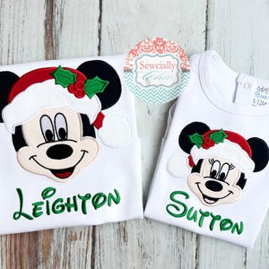 May include: Two white Christmas-themed shirts featuring Mickey and Minnie Mouse. Each shirt has a smiling character face wearing a Santa hat with holly. The name "Leighton" is embroidered in green on one shirt, and "Sutton" on the other.