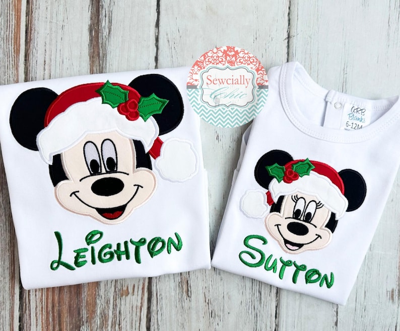 May include: Two white Christmas-themed shirts featuring Mickey and Minnie Mouse. Each shirt has a smiling character face wearing a Santa hat with holly. The name "Leighton" is embroidered in green on one shirt, and "Sutton" on the other.