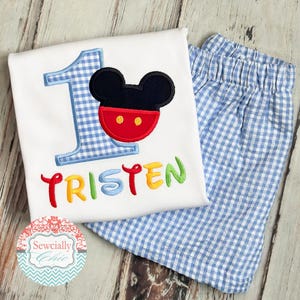 May include: A white toddler's birthday outfit featuring a Mickey Mouse design. The shirt has a blue gingham number 1, a Mickey Mouse face, and the name "Tristen" in colorful embroidery. The outfit includes matching blue and white gingham shorts.