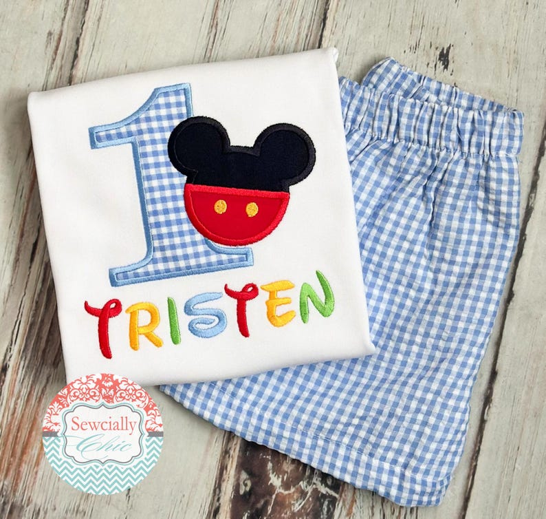 May include: A white toddler's birthday outfit featuring a Mickey Mouse design. The shirt has a blue gingham number 1, a Mickey Mouse face, and the name "Tristen" in colorful embroidery. The outfit includes matching blue and white gingham shorts.