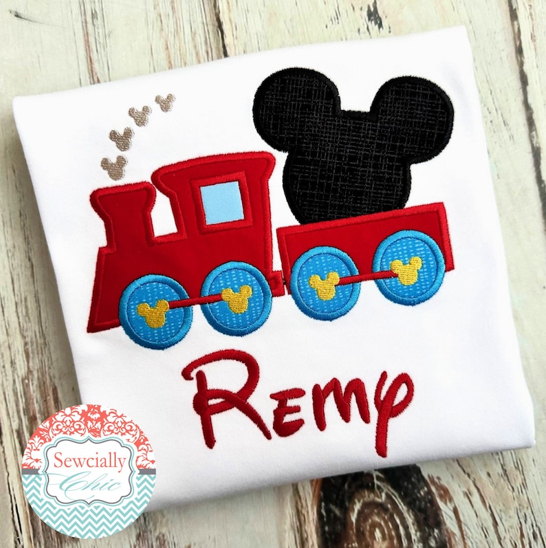 May include: A white t-shirt with a red and blue embroidered train with Mickey Mouse ears on top. The train has yellow wheels with Mickey Mouse ears on them. The name "Remy" is embroidered in red below the train.