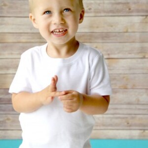 May include: A young boy wearing a white t-shirt with a crew neck and short sleeves. He is smiling and looking at the camera. The background is a wood-paneled wall.