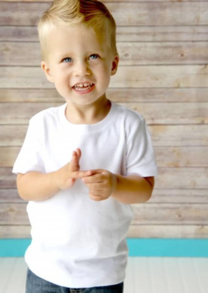 May include: A young boy wearing a white t-shirt with a crew neck and short sleeves. He is smiling and looking at the camera. The background is a wood-paneled wall.