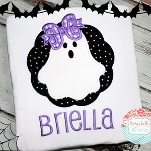 May include: White t-shirt with a purple bow and a ghost with black polka dots. The name "Briella" is embroidered in purple below the ghost. The t-shirt is on a wooden background with a spiderweb in the corner.