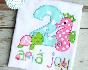 Puede incluir: Una camiseta blanca con un número dos de lunares azules y blancos, un caballito de mar de lunares rosas y blancos, una tortuga verde y rosa, y el nombre "Aria Joy" bordado en verde.