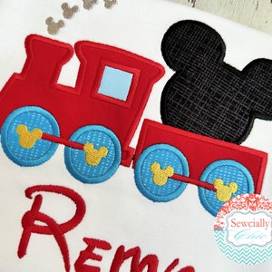 May include: Red and blue embroidered train with a black Mickey Mouse head on top. The train has yellow Mickey Mouse ears on each wheel. The name "Remy" is embroidered below the train.