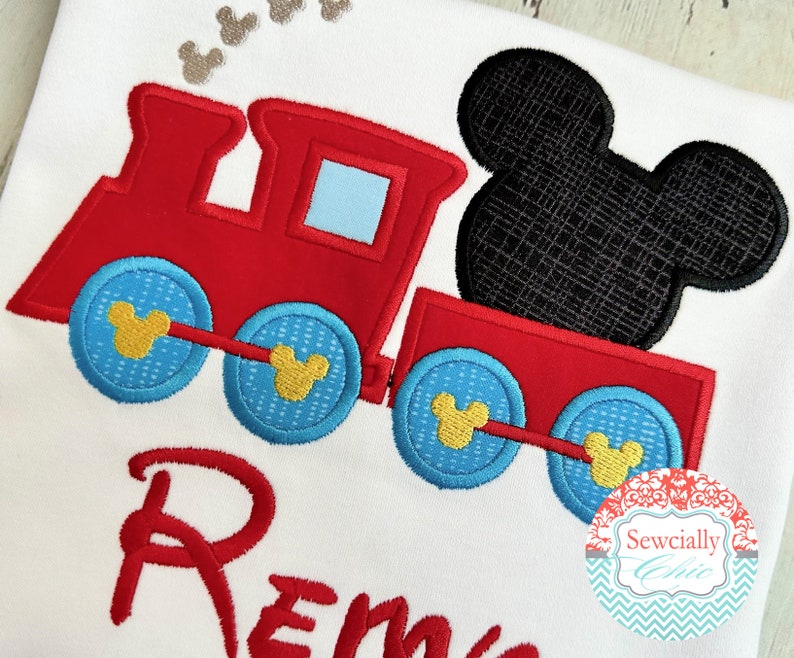 May include: Red and blue embroidered train with a black Mickey Mouse head on top. The train has yellow Mickey Mouse ears on each wheel. The name "Remy" is embroidered below the train.