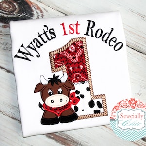 K&ouml;nnte beinhalten: Wei&szlig;es T-Shirt mit gesticktem Text "Wyatt's 1st Rodeo" und einem braunen und schwarzen Cartoon-Stier mit rotem Halstuch. Die Zahl 1 ist aus einem roten Bandana-Muster, schwarz-wei&szlig;em Kuhmuster und braunem Seil gefertigt.