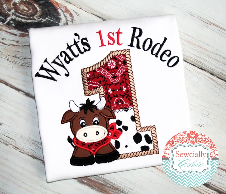 K&ouml;nnte beinhalten: Wei&szlig;es T-Shirt mit gesticktem Text "Wyatt's 1st Rodeo" und einem braunen und schwarzen Cartoon-Stier mit rotem Halstuch. Die Zahl 1 ist aus einem roten Bandana-Muster, schwarz-wei&szlig;em Kuhmuster und braunem Seil gefertigt.