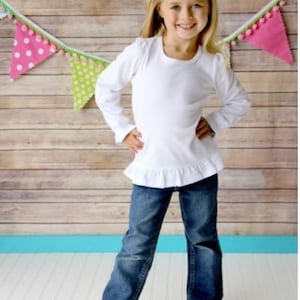 May include: A young girl wearing a white long-sleeved shirt with a ruffle hem and blue jeans. She is wearing a white bow in her hair. The girl is standing in front of a wooden wall with a colorful banner hanging above her.