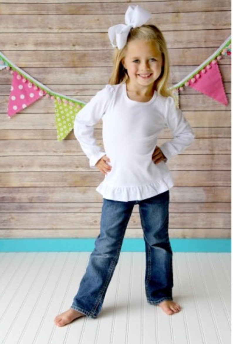 May include: A young girl wearing a white long-sleeved shirt with a ruffle hem and blue jeans. She is wearing a white bow in her hair. The girl is standing in front of a wooden wall with a colorful banner hanging above her.