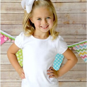 May include: A young girl wearing a white t-shirt with a large white bow in her hair. She is standing in front of a wooden background with colorful flags.