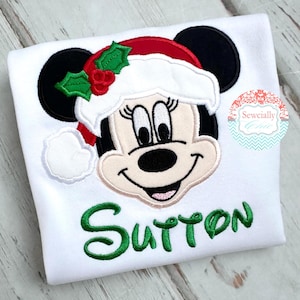 May include: White t-shirt featuring a Minnie Mouse design wearing a Santa hat with holly accents. The name "Sutton" is embroidered in green script below the character. The design is centered on the shirt.