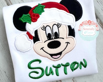 May include: White t-shirt featuring a Minnie Mouse design wearing a Santa hat with holly accents. The name "Sutton" is embroidered in green script below the character. The design is centered on the shirt.