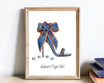 May include: A framed watercolor print featuring a blue high-heeled sandal with an orange-trimmed bow at the heel, accompanied by small blue paw prints on the floor and the text Auburn Tiger Girl printed in black at the bottom of the white page.