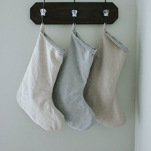 May include: Three linen stockings hanging on hooks, one is beige, one is gray, and one is white. The stockings have a black and white striped trim.