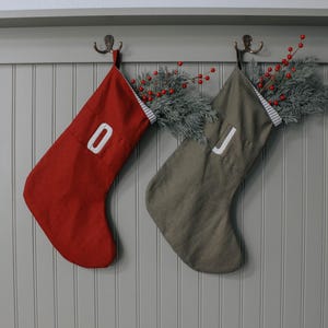 May include: Two Christmas stockings hang from hooks on a gray wall. One stocking is red with a white "0", and the other is olive green with a white "J". Both are filled with faux greenery and red berries.