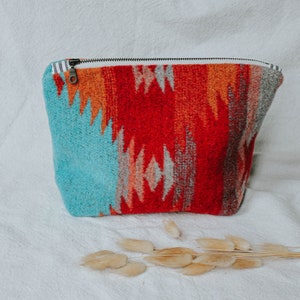 Pode incluir: Uma bolsa tecida colorida com fecho de correr. A bolsa apresenta um padrão vibrante em turquesa, vermelho, laranja e cinza. O fecho de correr é preto com um puxador de cor bronze. O forro interior é branco com uma risca a preto e branco.