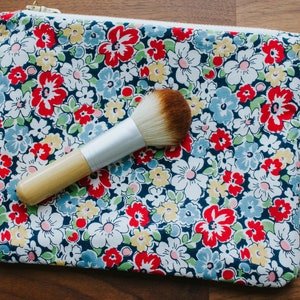 May include: A blue floral makeup bag with a zipper closure and a white label with the text "Ha!" on it. A wooden makeup brush with a silver handle is inside the bag.