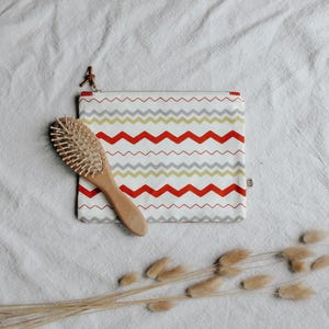 May include: A wooden hairbrush with a light brown handle and a rectangular pouch with a zipper. The pouch has a white background with a red, gray, and yellow zigzag pattern. The zipper pull is brown leather.