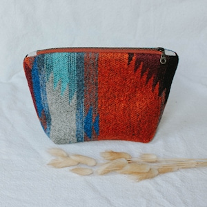 May include: A small, zippered pouch with a colorful, geometric pattern in shades of red, blue, and gray. The pouch is made of a woven fabric and has a brown zipper.