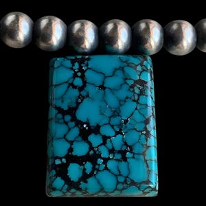 May include: A rectangular turquoise pendant with a striking matrix pattern of black veins. The pendant is paired with a strand of silver-colored, round beads, creating a bold contrast against the black background.