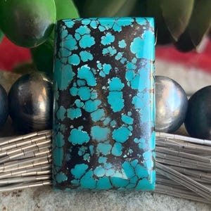 May include: A rectangular turquoise stone with a striking pattern of blue and black. The stone is set against a backdrop of silver beads and wire, with green foliage in the background. The turquoise stone is the focal point of a piece of jewelry.