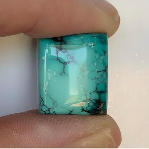 May include: A rectangular turquoise gemstone with a polished surface. The stone has a vibrant teal color with dark, web-like veins running throughout. The gemstone is held between two fingers.