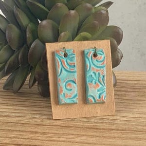 May include: A pair of turquoise blue ceramic earrings with a copper-toned floral pattern. The earrings are rectangular in shape and have a simple wire hook.
