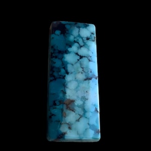 May include: A rectangular turquoise gemstone with a polished surface. The stone features a mottled pattern of light and dark blue hues, with brown veining. The gemstone is set against a black background.