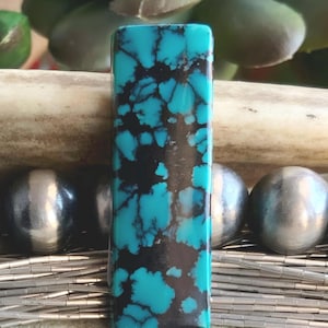 May include: A rectangular turquoise gemstone with a striking black matrix pattern. The polished stone displays a vibrant blue-green color, with dark, irregular veins creating a unique design. The gemstone is set against a blurred background.