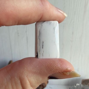 May include: A rectangular white gemstone cabochon featuring smooth, polished surfaces and distinct dark grey to black veining patterns running vertically across the stone, showcasing a natural mineral texture with a glossy, reflective finish.