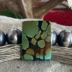 May include: A rectangular turquoise gemstone with a mottled pattern of green, brown, and black. The stone is set between silver beads and a strand of silver tubes, suggesting a jewelry design. The background is a neutral, textured surface.