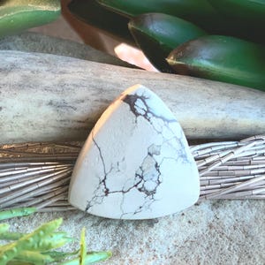 May include: A triangular, off-white gemstone with dark brown veining. The stone is set against a backdrop of silver beads, a weathered wooden piece, and green foliage. The stone's shape is reminiscent of a shield or a pendant.