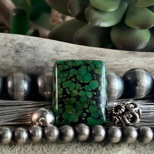 May include: A rectangular green turquoise cabochon with a black matrix pattern, positioned among a collection of smooth, round silver beads and a single ornate, textured silver bead, creating a detailed jewelry component arrangement.