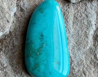 May include: A smooth, polished turquoise cabochon with a light blue-green color and a mottled pattern. The stone is oval-shaped and has a glossy finish.