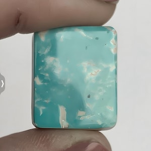 May include: A rectangular turquoise gemstone with a polished surface. The stone features a marbled pattern of white and light brown inclusions, creating a unique visual texture. The gemstone is held between two fingers.