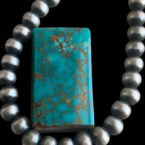 May include: A necklace featuring a rectangular turquoise pendant with brown matrix veining, suspended from a strand of round, polished silver-toned beads. The turquoise stone displays a vibrant blue-green hue with intricate, web-like brown patterns.