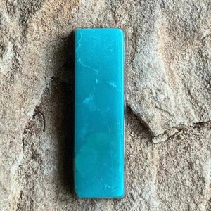 May include: A rectangular, turquoise-colored stone with white veining. The polished stone is set against a rough, tan-colored surface. The stone's shape is a long rectangle.