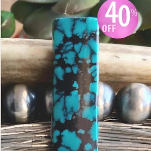 May include: A rectangular turquoise stone with a vibrant blue color and dark black matrix webbing. The stone is positioned vertically in front of several polished silver beads and thin silver tubes. A pink circular graphic displays 40% OFF.