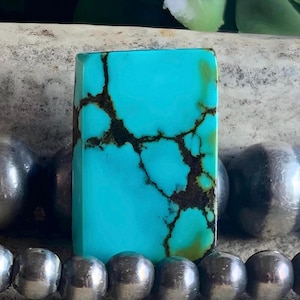 May include: A rectangular turquoise gemstone with dark brown matrix veining. The polished stone is a vibrant blue-green color. The gemstone is displayed with a strand of silver beads in the foreground, suggesting jewelry or craft use.