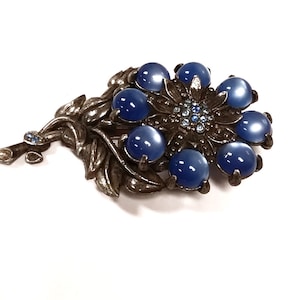 Puede incluir: Un broche de tono plateado con un diseño de flor. La flor tiene ocho piedras de cabujón azules y un centro de piedras más pequeñas transparentes. La flor está unida a un tallo de tono plateado con hojas.