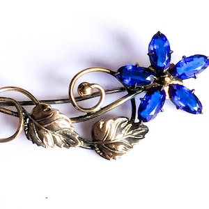 Puede incluir: Broche vintage con diseño floral y pétalos de gemas azules. El broche tiene un tallo de metal dorado con hojas y espirales decorativas, creando un elegante accesorio inspirado en la naturaleza.