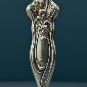 May include: A silver-toned metal handle featuring an embossed floral design with flowing, curved lines and organic shapes. The surface has a polished, reflective finish with intricate relief details of flowers and stems extending along the length.