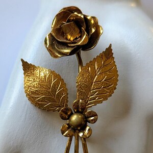May include: A gold-toned metal brooch in the shape of a rose with leaves and a small flower at the base.