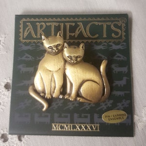May include: A gold-toned metal pin featuring two cats sitting together. The pin is on a green card with the word "ARTIFACTS" in gold lettering and the date "MCMILXXXVI".