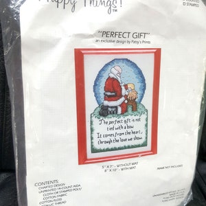 Peut inclure: Un kit de point de croix représentant un Père Noël et un enfant dans un globe de neige. Le kit comprend un tissu Aida 14 points, du fil à broder en coton, du fil métallique, une aiguille et des instructions. Le kit mesure 13 cm par 18 cm sans passe-partout et 20 cm par 25 cm avec passe-partout. Le texte sur le kit est "Le cadeau parfait n'est pas attaché avec un nœud. Il vient du cœur, à travers l'amour que nous montrons."